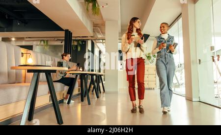 Junge Geschäftsfrauen, die morgens durch ein modernes Büro laufen. Zwei junge Geschäftsfrauen halten Kaffeetassen in der Hand und unterhalten sich. Unternehmerin Stockfoto