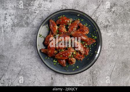 Honigglasierte Chicken Wings mit Sesam und Kräutern auf einem runden Teller auf dunkelgrauem Hintergrund. Draufsicht, flach liegend Stockfoto