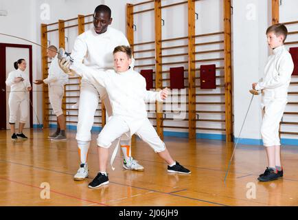 Fokussierte fleißig seriöse positive Jungen fechten aufmerksam dem professionellen freundlichen Fechttrainer im Fitnessstudio zu Stockfoto