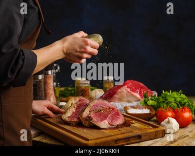 Rohes Rindfleisch, Schweinefleisch, Kalbssteaks, zubereitet von den Händen des Küchenchefs auf einem Holztisch, dunkelblauer Hintergrund. Das Konzept ist Restaurant und hausgemachte Rezepte. Werbung Stockfoto