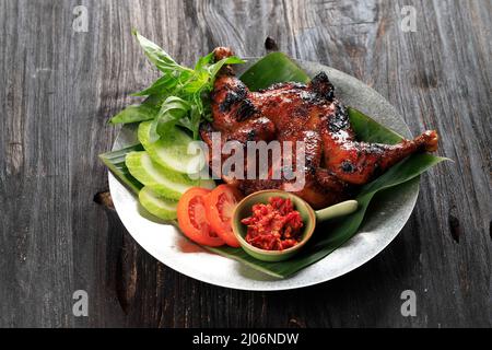 Ayam Bakar Bakakak oder ganz gebratenes Huhn mit süßer Sojasauce, serviert mit Gurke, Tomate und Basilikum. Indonesian Popular Dish Geflügel Stockfoto