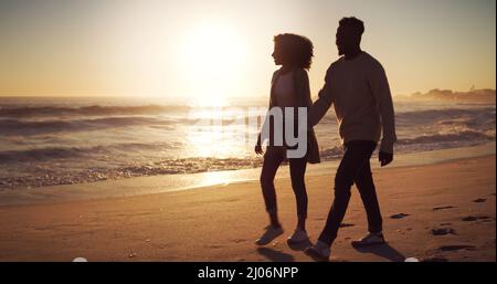 Seite an Seite auf jedem Lebensweg. Ganzkörperaufnahme eines liebevollen jungen Paares, das bei Sonnenuntergang am Strand spazieren geht. Stockfoto