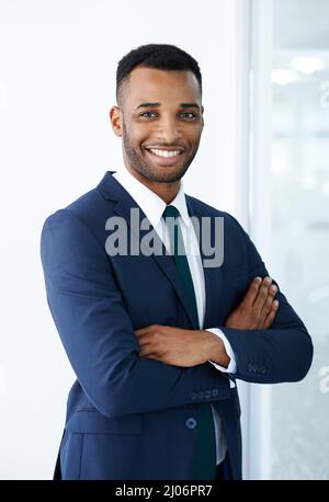 Selbstbewusster Charme für Unternehmen. Ein hübscher junger afroamerikanischer Geschäftsmann, der mit gefalteten Armen drinnen steht. Stockfoto