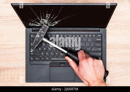 Der Laptop-Bildschirm ist mit einem Hammer von der Hand gebrochen. Konzept der groben Reparatur der Computerausrüstung. Stockfoto