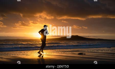 Silhouette Mann läuft am Milford Strand bei Sonnenaufgang, Rangitoto Island im Hintergrund, Auckland. Stockfoto