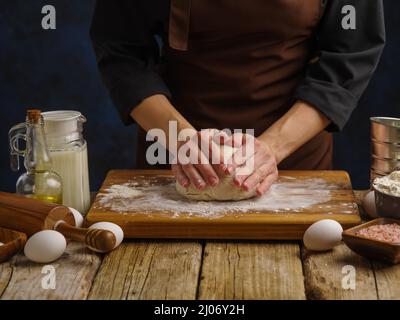 Ein professioneller Koch knetet den Teig auf einem bemehlten Holzschneidebrett auf einem dunkelblauen Hintergrund. Rezepte für Brot, Pizza, Pasta, Kuchen, Spaghetti. Piz Stockfoto