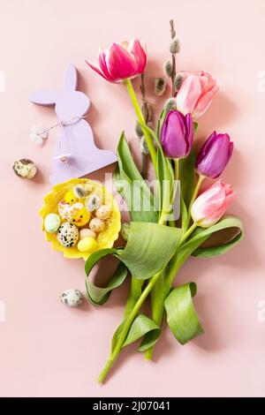 Rosa und lila Tulpen mit bunten Wachteleiern in einem Nest auf rosa Hintergrund. Konzept für Frühlings- und Osterferien. Draufsicht flach liegend. Stockfoto