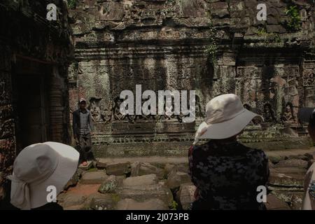Frauen, die stehen, während sie einem Reiseleiter zuhören, der Ihnen etwas erklärt, und überblicken eine verwitterte Wand, die mit Basreliefs dekoriert ist, im Preah Khan Tempel in Siem Reap, Kambodscha. Stockfoto