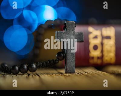 Ein Kreuz und ein Rosenkranz vor dem Hintergrund einer heiligen bibel auf einem Holztisch in einer Kirche mit Lichtern.das Konzept von Gebet und Glauben i Stockfoto