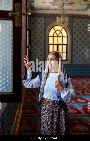 Eine wunderschöne ukrainische Frau mit einem Schal auf dem Kopf steht in der Moschee am Fenster und betet für den Frieden. Stockfoto