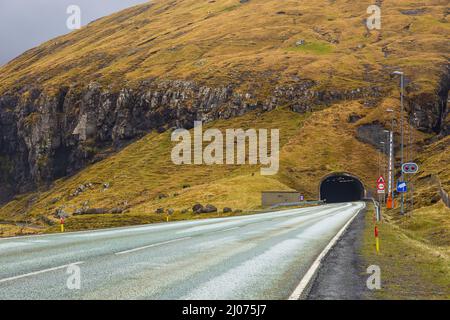Gaschadalur, Färöer Inseln - 04. Mai 2018: Asphaltstraße entlang der Hügel in Gaschadalur auf der Insel Vagar. Tunnel in den Fels gehauen. Kleines Dorf liegt o Stockfoto