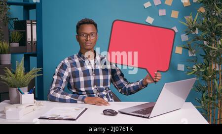Junger Erwachsener, der die Kamera anschaut und Sprechblase für Textnachrichten hält und leere Kartentafel zeigt. Person, die eine Kommunikationsvorlage in einem Karton erstellt. Stockfoto