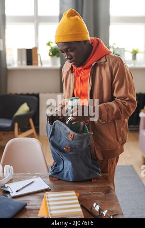 Vertikale, mittellange Aufnahme eines jungen Studenten der amerikanischen Universität Frican, der am Schreibtisch stand und eine Flasche Wasser in den Rucksack legte Stockfoto