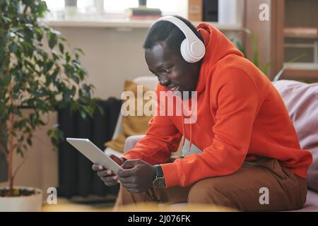 Fröhlicher afroamerikanischer Kerl mit leuchtend orangefarbenem Hoodie und weißen Kopfhörern, der mit Freunden über ein digitales Tablet chattet und Platz zum Kopieren verwendet Stockfoto