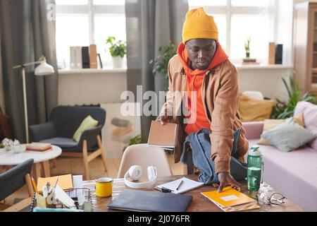 Horizontale Aufnahme eines modernen afroamerikanischen College-Studenten, der am Schreibtisch steht und sich auf die Schule vorbereitet, indem er Lehrbücher und Copybooks in den Rucksack packt Stockfoto