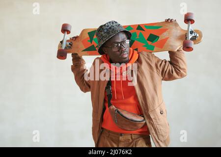 Mittleres Studioporträt eines hübschen jungen schwarzen Mannes in stilvollem Casual-Outfit und einer Brille, die Skateboard hinter dem Kopf hält und wegschaut Stockfoto