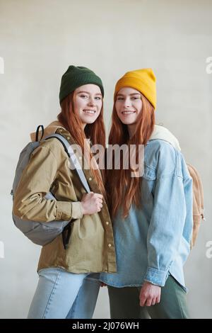 Vertikales mittellange Studioporträt von zwei rothaarigen jungen kaukasischen Zwillingen, die stilvolle Casual-Outfits tragen und Rucksäcke lächeln an der Kamera Stockfoto