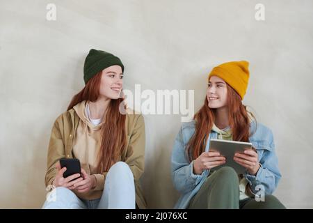 Studio Portrait von zwei schönen jungen Schwestern in lässiger Kleidung sitzen zusammen auf dem Boden Surfen Interent auf Gadgets Blick auf einander Stockfoto