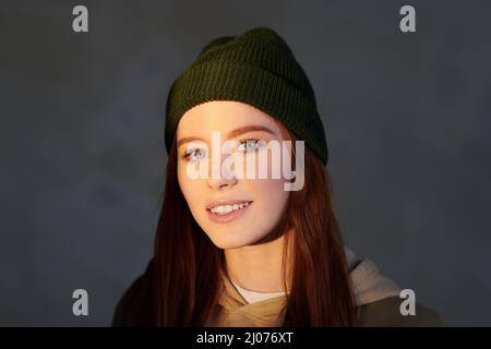 Horizonatale Nahaufnahme einer attraktiven jungen Frau mit dunkelrotem Haar und Mütze, die die Kamera anlächelt, grauem Hintergrund Stockfoto