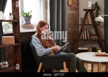 Horizontale mittlere lange Seitenansicht Aufnahme der schönen Frau mit langen roten Haaren sitzen entspannt im Stuhl im Wohnzimmer Surfen Interent auf digitalem Tablet Stockfoto