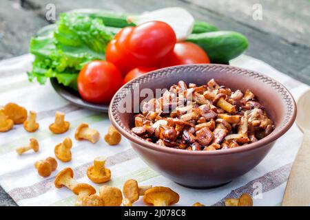 Gebratene Waldpilze Pfifferlinge mit Zwiebel in rustikaler Schüssel und Teller mit frischem Gemüse für Salat auf dem Hintergrund Stockfoto