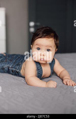 Porträt eines traurigen Babys, das Denim-Strampler trägt und zu Hause auf einer grauen Decke liegt. Aufgewühlt aussehendes Kleinkind kriecht im Schlafzimmer auf dem Bett, erkundet die Welt. Selektiv Stockfoto