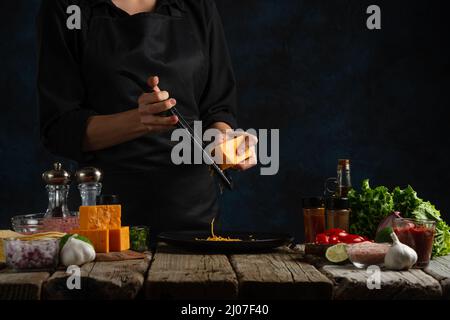 Professioneller Koch in schwarzer Uniform rostet Käse auf den Teller. Backstage mit traditionellen mexikanischen Tacos auf dunkelblauem Hintergrund. Eingefrorene Bewegung. Stockfoto