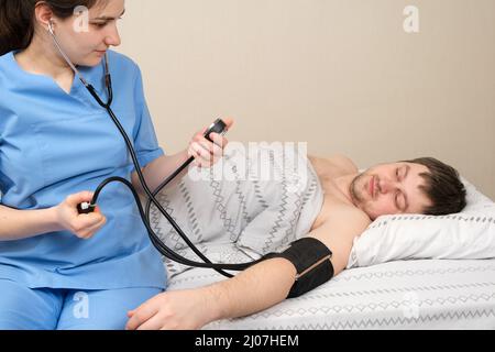 Der Arzt misst den Blutdruck eines jungen 30-jährigen Mannes, der im Bett liegt. Erkrankungen des Herzens und der Blutgefäße beim Menschen, mechanisch Stockfoto
