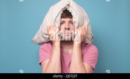 Der kaukasische junge Mann ist müde und sehr müde, wenn er sein Kopfkissen auf dem Kopf hält. Stockfoto