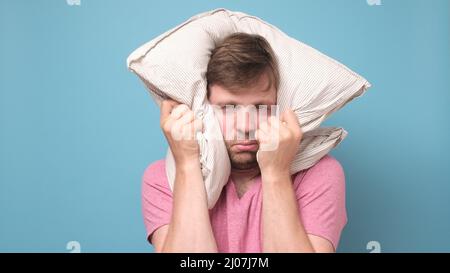 Der kaukasische junge Mann ist müde und sehr müde, wenn er sein Kopfkissen auf dem Kopf hält. Stockfoto