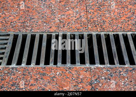 Drainage-Gitter mit Eisenstreifen auf dem Gehweg mit einer Oberfläche aus rotem Granit, Nahaufnahme eines Sturmsystems mit großem Loch. Stockfoto