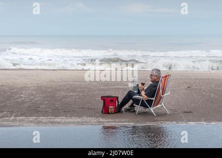 Reifer Mann mit grauen Haaren und Gläsern auf einem Strandstuhl sitzend, die sich alle im Wasser und im Hintergrund die Wellen des Meeres widerspiegeln. Stockfoto