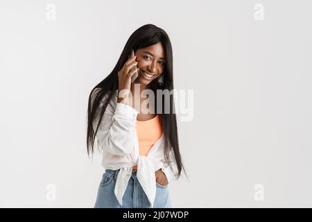 Lächelnde junge afrikanische Frau mit langen, dunklen Haaren, die über einem weißen Wandhintergrund stehen und das Mobiltelefon halten Stockfoto