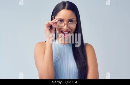 Oh, Wow. Ein beschnittenes Porträt einer attraktiven jungen Frau, die im Studio vor grauem Hintergrund über ihre Brille gupft. Stockfoto