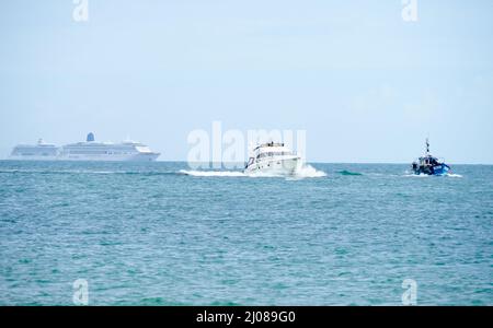 Ein kleines Fischerboot und ein Motorcruiser mit zwei großen Kreuzfahrtschiffen, die im Hintergrund auf See festgemacht sind Stockfoto