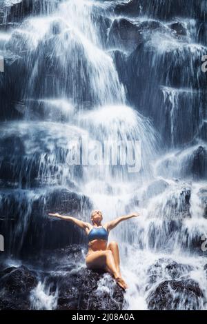 Reisen Sie im Bali-Dschungel. Schöne junge Frau, die unter dem fallenden Quellwasser auf Felsen sitzt, genießt einen tropischen Wasserfall. Asiatische Natur, Tagesausflug Stockfoto
