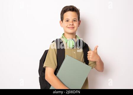 Junge trägt Kopfhörer und Rucksack hält ein Buch über isolierten Hintergrund glücklich tun okay Zeichen, Daumen nach oben mit dem Finger, ausgezeichnete Zeichen. Stockfoto