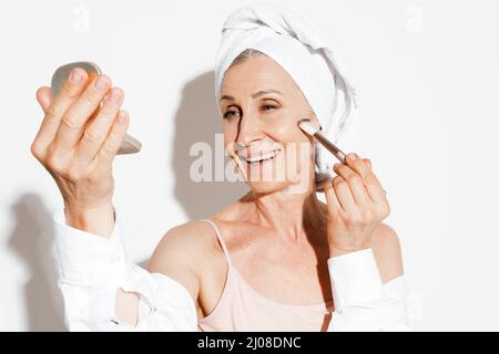 Ältere Frau in ein Handtuch gehüllt lächelnd, während Make-up isoliert über weiße Wand Stockfoto