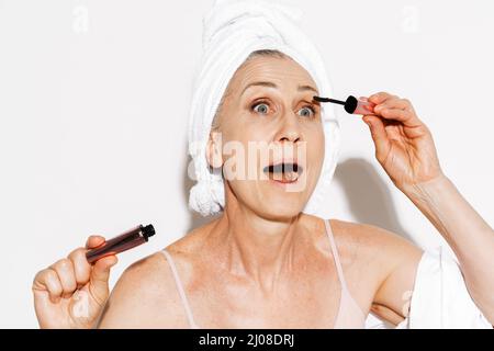 Ältere Frau in ein Handtuch eingewickelt, das Mascara aufgetragen, während sie sich isoliert über die weiße Wand schminkt Stockfoto