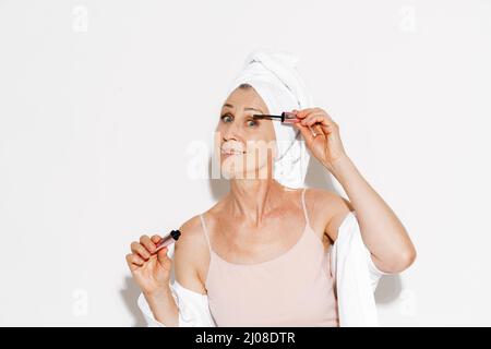 Ältere Frau in ein Handtuch eingewickelt, das Mascara aufgetragen, während sie sich isoliert über die weiße Wand schminkt Stockfoto