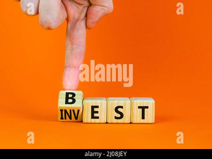 Symbol für beste Investition. Geschäftsmann dreht einen Holzwürfel und ändert das Wort Invest in Best. Schöner orangefarbener Tisch, orangefarbener Hintergrund. Business und Best Stockfoto