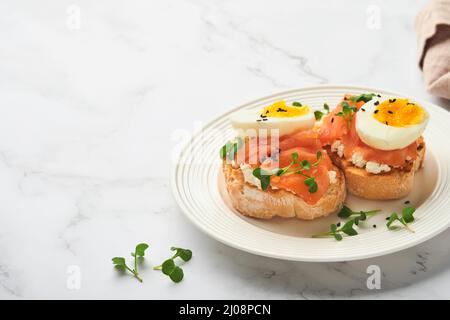 Sandwich mit köstlichen Belägen, geräuchertem Lachs, Eiern, Kräutern und Mikrogrüns Rettich, schwarzen Sesam über weißem Teller auf weißem Marmortisch Backgro Stockfoto