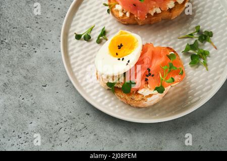 Sandwich mit leckeren Belägen geräucherter Lachs, Eier, Kräuter und Mikrogrüns Rettich, schwarze Sesam-Samen über weißem Teller auf grauem Beton Tisch Backgro Stockfoto