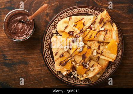 Crepes-Suzette mit Orangen und Schokolade. Dünne Crepes mit Schokoladenaufstrich, Haselnüssen und Orangenscheiben Obst auf dem Teller zum Frühstück auf altem dunklen Woo Stockfoto