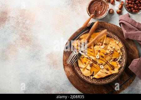 Crepes-Suzette mit Orangen und Schokolade. Dünne Crepes mit Schokoladenaufstrich, Haselnüssen und Orangenscheiben Obst auf dem Teller zum Frühstück auf altem Beton Stockfoto