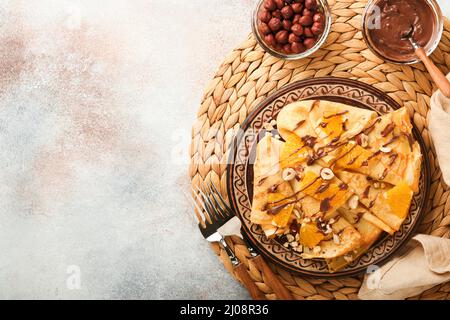 Crepes-Suzette mit Orangen und Schokolade. Dünne Crepes mit Schokoladenaufstrich, Haselnüssen und Orangenscheiben Obst auf dem Teller zum Frühstück auf altem Beton Stockfoto