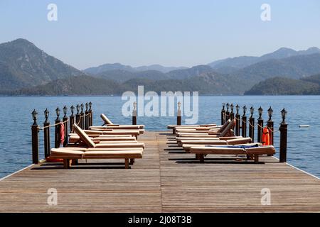 Strandurlaub am Meer Resort in der Ägäis Küste der Türkei, Liegestühle auf einem hölzernen Pier gegen Berge. Leere Sonnenliegen auf grünem Uferhintergrund Stockfoto