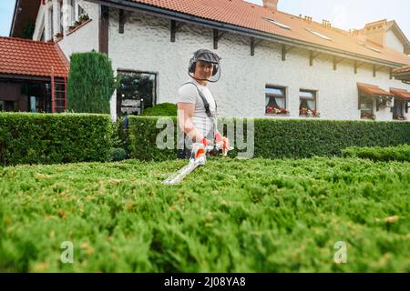 Hübscher junger Mann in Arbeitskleidung, Schutzbrille und Handschuhe, die im Sommer grüne Sträucher im Garten beschneiden. Konzept der saisonalen Arbeit und Landschaftsgestaltung. Stockfoto