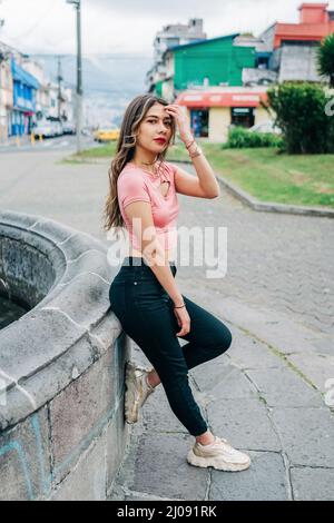 Ganzer Körper von attraktiven jungen lateinischen Frau, die sich an einer Steinwand lehnt, Make-up, Freizeitkleidung, lange blonde Haare, Stadtleben trägt Stockfoto