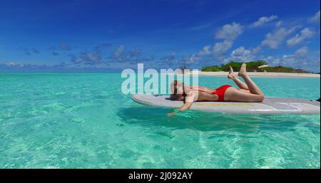 Kaukasische blonde Frau in einem roten Badeanzug, die auf dem Surfbrett im Ozean auf den Malediven liegt Stockfoto